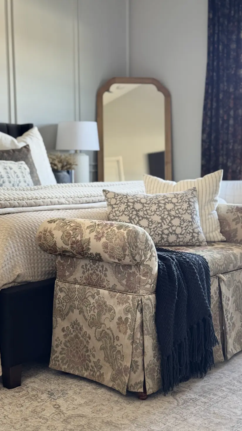 A floral-upholstered armchair with a navy throw, layered patterned pillows, and a wood-framed mirror