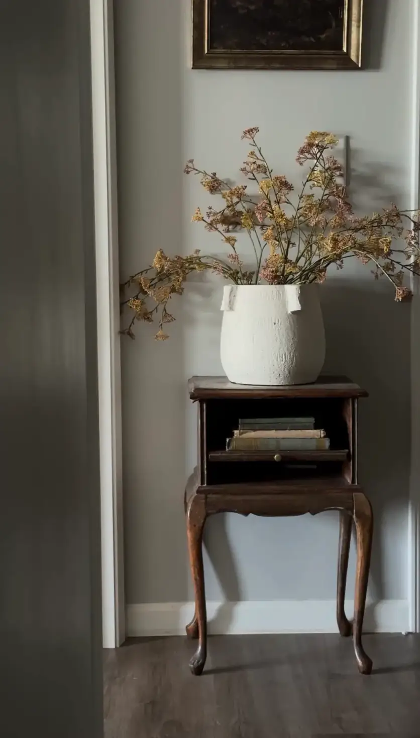 An antique wood side table holding a white vase of dried flowers and a short stack of books