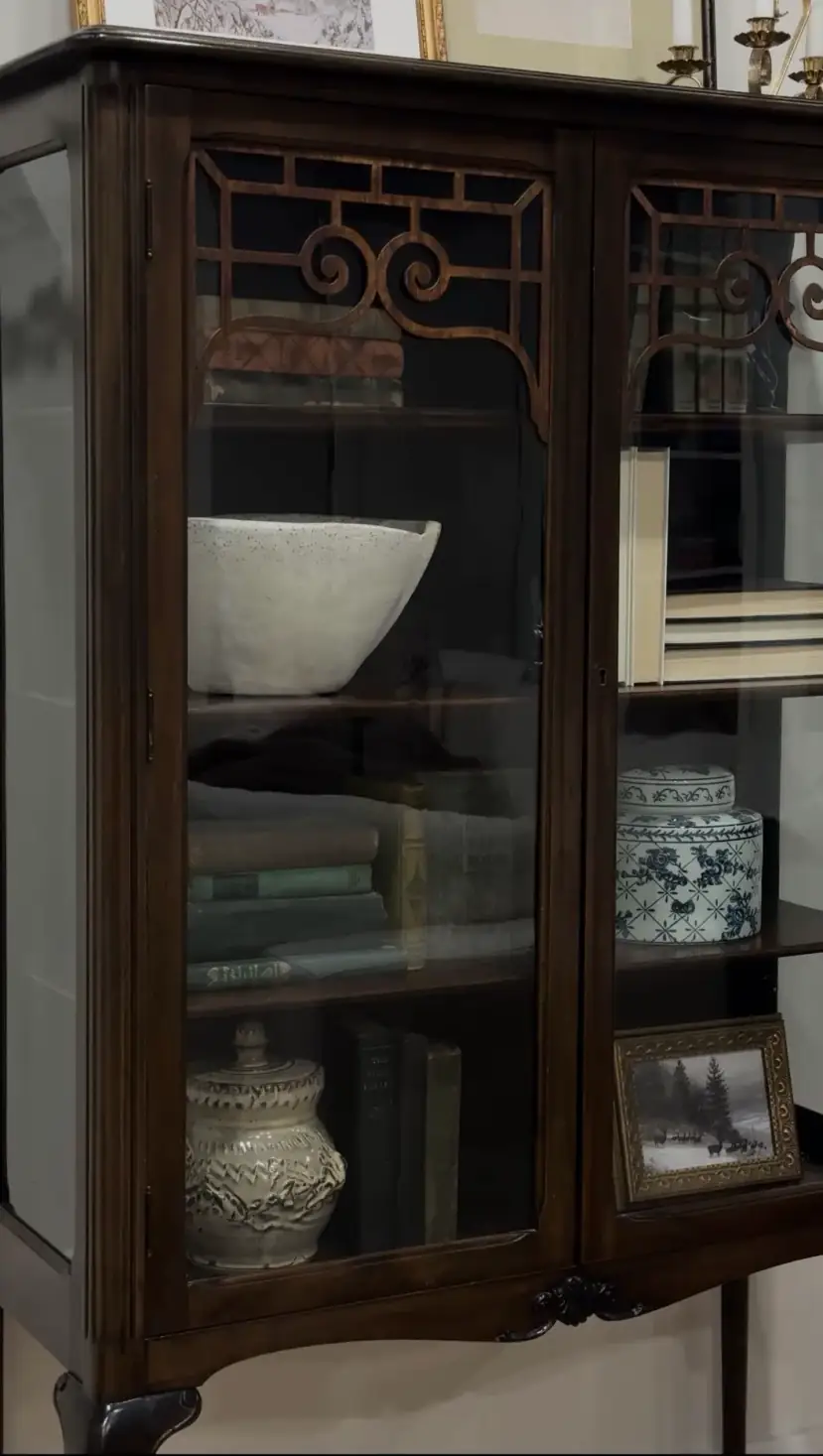 A styled glass cabinet with pottery, stacked books, and a winter landscape in a small frame