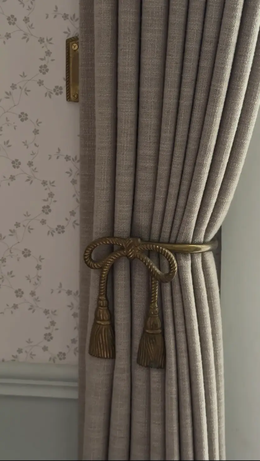 A gray curtain held by a brass bow tieback against floral wallpaper