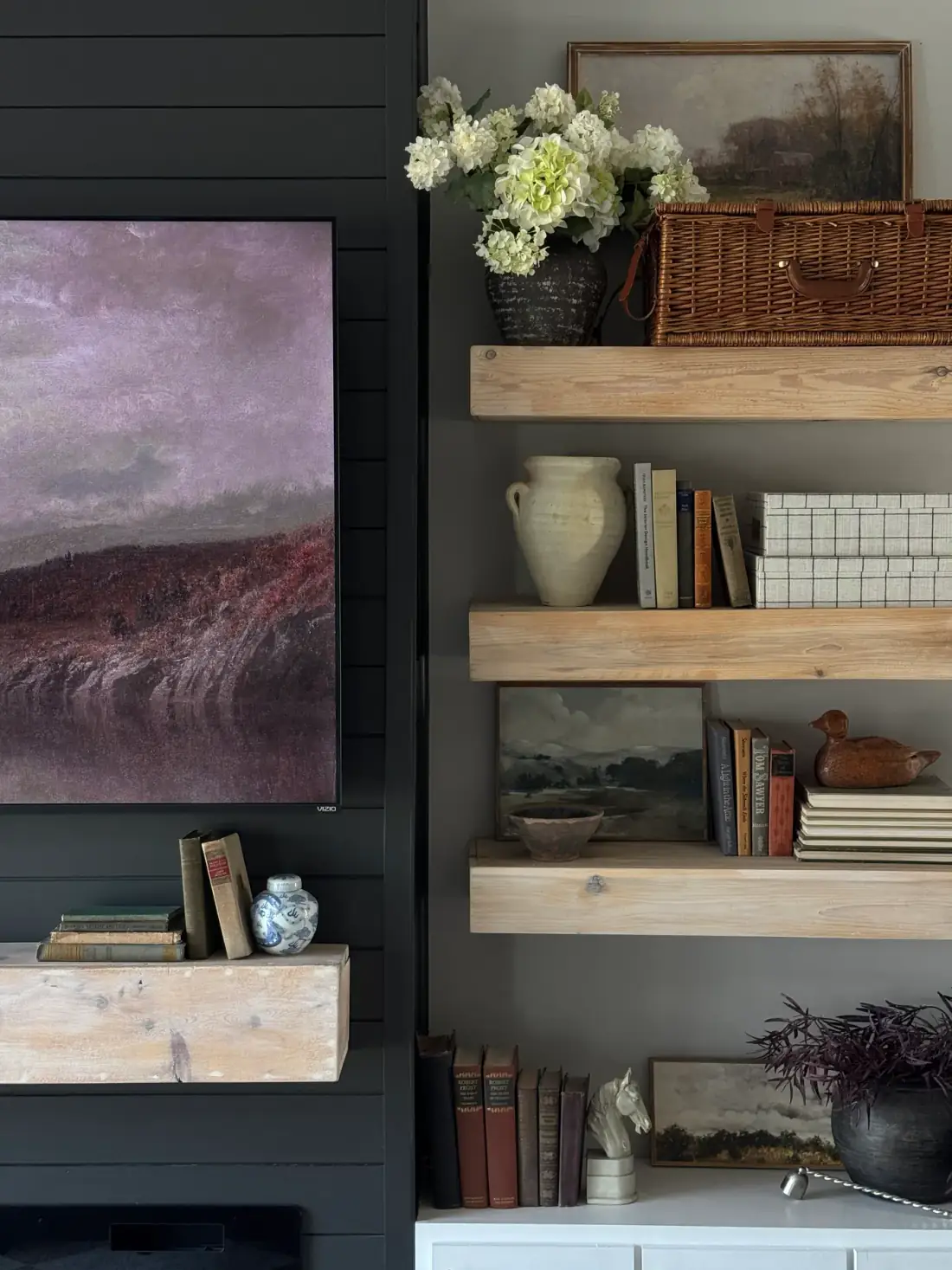 Oak shelves with hydrangeas, a woven basket, stacked books, a wooden duck, and a landscape on the black wall